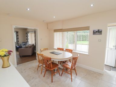 A dining area with a round table and six wooden chairs next to a window with vertical blinds and an open door at Awel Y Berwyn in Llangynog