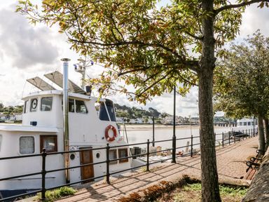 A boat docked along a riverside walkway with trees benches and houses across the river at Stonewood Lodge in Woolsery