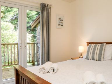 A bedroom with a wooden bed a striped pillow rolled towels a lamp on a side table and glass doors leading to a balcony at Stonewood Lodge in Woolsery