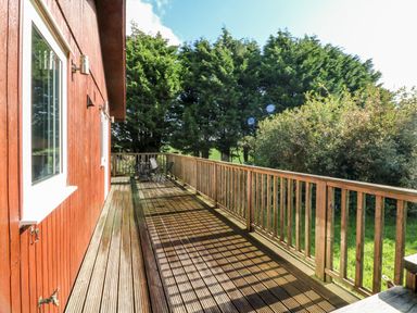 A wooden deck attached to a red house with outdoor chairs and a table surrounded by trees and greenery at Stonewood Lodge in Woolsery