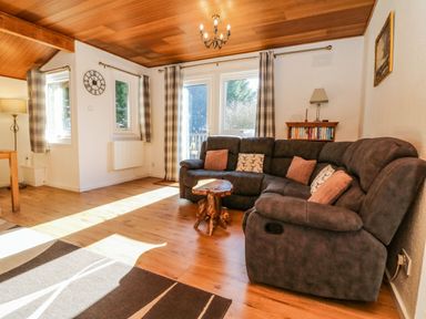 A living room with a large sectional sofa, small wooden table, bookshelves, and windows with plaid curtains at Stonewood Lodge in Woolsery