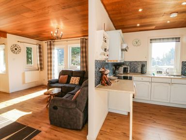 An open living room with grey sofas and a wooden coffee table next to a kitchen with white cabinets and a countertop at Stonewood Lodge in Woolsery