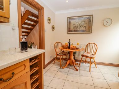 A dining area with a round wooden table and two chairs with cushions a bottle of wine and glasses on the table a wooden cabinet with a decanter and glasses on the countertop stairs visible through a doorway clock on the wall at Corner House in Cinderford