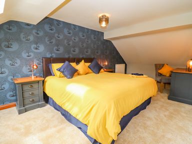 A bedroom with a large bed with yellow and blue pillows two side tables with lamps a chair with a cushion and wallpaper with tree patterns at Corner House in Cinderford