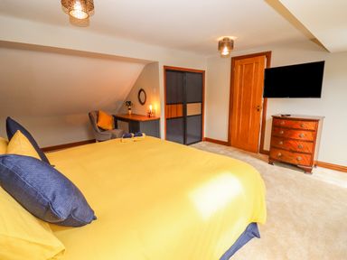 A bedroom with a yellow bedspread navy pillows a wooden dresser a wall-mounted television a desk with a chair and a door at Corner House in Cinderford