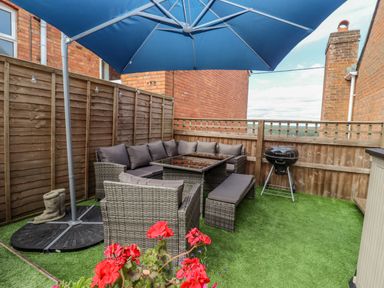 An outdoor patio area with rattan-style seating, a glass table, a bench, a blue umbrella, a barbecue grill, and artificial grass at Corner House in Cinderford