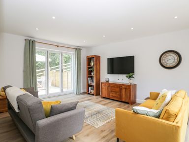 A living room with sofas a wall-mounted TV a wooden cabinet bookshelf and a wall clock at Stag Cottage in Lytchett Matravers