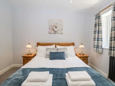 A bedroom with a bed and pillows two bedside tables with lamps and a window with checked curtains at Stag Cottage in Lytchett Matravers