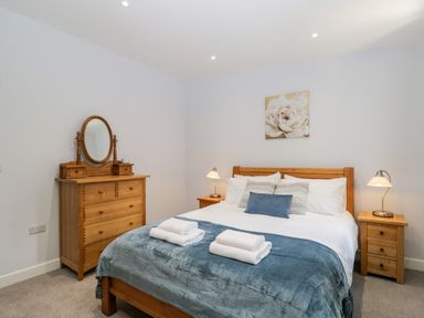 A bedroom with a wooden bed with blue and white bedding two bedside tables with lamps and a wooden dresser with an oval mirror at Stag Cottage in Lytchett Matravers