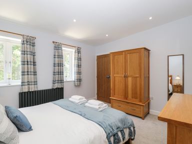 A bedroom with a bed folded towels a wooden wardrobe a wooden dresser and two windows with plaid curtains at Stag Cottage in Lytchett Matravers