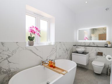 A bathroom with a freestanding bathtub and wooden bath tray with bottles a wall mounted sink and mirror and a toilet at Stag Cottage in Lytchett Matravers
