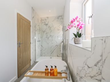 A bathroom with a bathtub, wooden door, shower area with glass door, and pink flowers on the windowsill at Stag Cottage in Lytchett Matravers