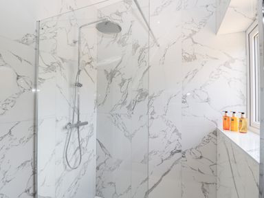 A shower area with a rainfall showerhead and a handheld shower on marble tiled walls with bottles on the windowsill at Stag Cottage in Lytchett Matravers