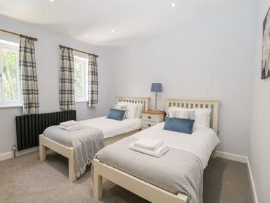 A bedroom with two single beds side by side a nightstand with a lamp between them and two windows with plaid curtains at Stag Cottage in Lytchett Matravers