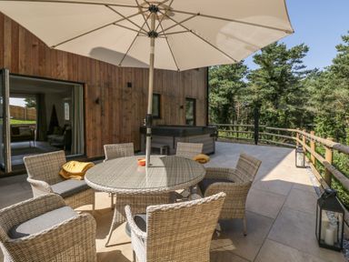 An outdoor patio with a round table and six chairs under an umbrella and a hot tub near a wooden building at Stag Cottage in Lytchett Matravers