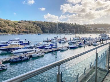 Boats docked at a marina with a glass railing in the foreground and wooded hills in the background at 15 Dart Marina in Dartmouth