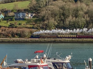 A steam train traveling along tracks by a river with boats in the foreground and houses on a hillside in the background at 15 Dart Marina in Dartmouth