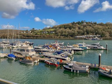 A marina with several boats and yachts docked on calm water with a hillside and houses in the background at 15 Dart Marina Dartmouth