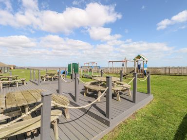 An outdoor seating area with wooden picnic tables and a children's playground behind a fenced grassy area at Bungalow 14 in Corton