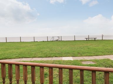 A grassy area with a wooden railing in the foreground a paved pathway a chain link fence a metal gate and a wooden bench at Bungalow 14 in Corton