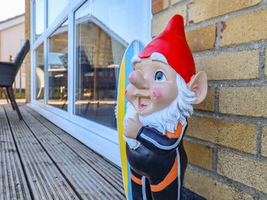 A garden gnome with a red hat and white beard leaning against a brick wall on a wooden deck outside a building with large windows at Bungalow 14 in Corton