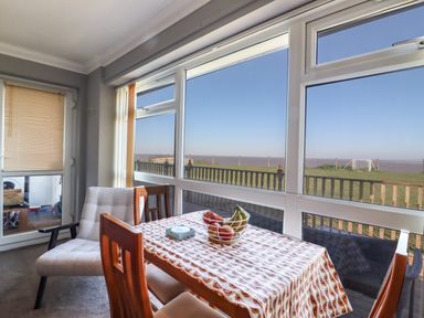 A dining area with a table covered by a tablecloth and a fruit bowl with chairs by large windows overlooking a grassy area and sea at Bungalow 14 in Corton