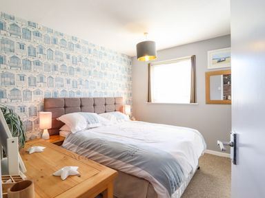 A bedroom with a double bed wooden bedside tables lamps a window with curtains and wallpaper with beach huts at Bungalow 14 in Corton