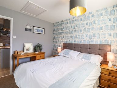 A bedroom with a bed side table lamps a wooden desk with a mirror and plant and wallpaper with blue houses at Bungalow 14 in Corton