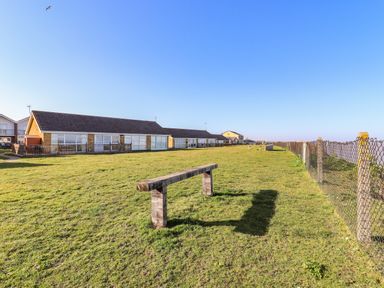 A grassy open area with a bench shed a shadow and single story residential buildings with large windows and fences at Bungalow 14 in Corton