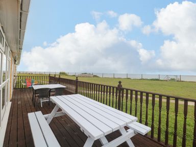 A wooden deck with white picnic tables and chairs overlooking a grassy area and the sea at Bungalow 14 in Corton
