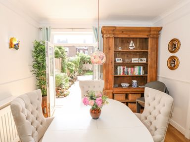 A dining room with a white table and four beige tufted chairs a wooden bookshelf with books and ornaments and garden view at Apple Tree House in Hunstanton