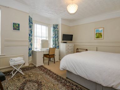 A bedroom with a bed, white dresser, TV, vanity desk with a chair, blue floral curtains, and a patterned rug at Apple Tree House in Hunstanton