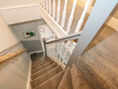 A carpeted staircase with white banisters and a wooden handrail leading down to a hallway with a patterned rug at Apple Tree House in Hunstanton