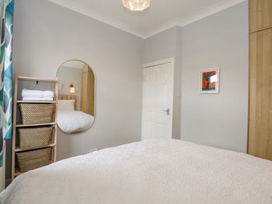 A bedroom with a white bedspread wooden shelves with baskets an oval mirror a white door and a framed picture on the wall at Apple Tree House in Hunstanton