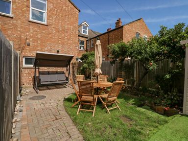 A backyard with a wooden table and chairs set on grass and a swing bench on a paved area at Apple Tree House in Hunstanton