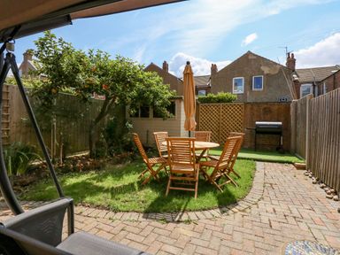 A backyard with a round wooden table and six chairs an umbrella a fruit tree a barbecue grill and a shed at Apple Tree House in Hunstanton