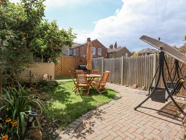 A garden with a wooden table and chairs an umbrella a swing a tree and a paved pathway at Apple Tree House in Hunstanton