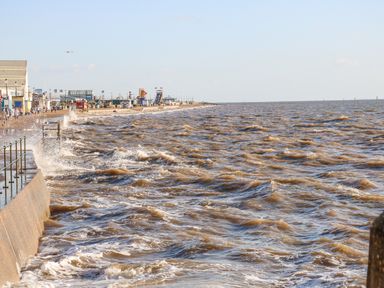 A seawall with waves crashing and a promenade with people and buildings in the distance at Apple Tree House in Hunstanton