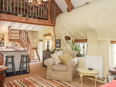 A living room with a beige armchair and pillows a small table and plants near windows and stairs at The Byre in Monkleigh near Great Torrington