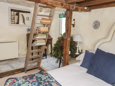A bedroom with a bed blue pillows a floral blanket a wooden ladder with books a wooden desk a plant and rustic wooden beams at The Byre in Monkleigh near Great Torrington