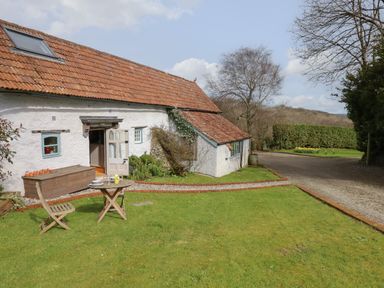 A garden with a wooden table and chair set outside a white cottage with a red tiled roof at The Byre in Monkleigh near Great Torrington