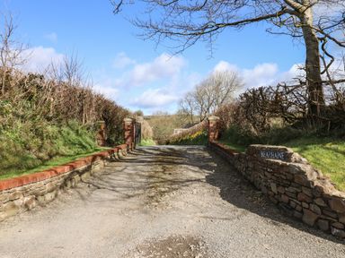 A gravel driveway with stone walls and brick pillars leading to a gate surrounded by hedges and trees at Bearhaine