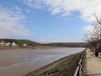 A river with a paved walkway and railing along the right side and houses and green hills on the left at The Byre in Monkleigh near Great Torrington