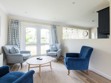 A living room with two blue armchairs two light grey armchairs a wooden coffee table and glass doors with curtains at Silverdale in Rock