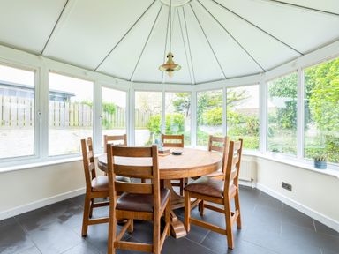 A dining area with a round wooden table and six wooden chairs in a sunroom with large windows at Silverdale in Rock