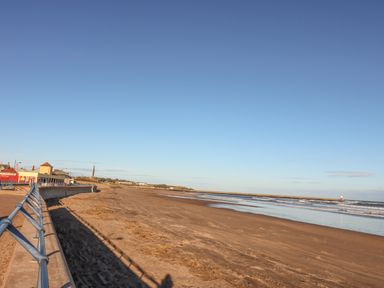 A sandy beach with a metal railing along a paved walkway and buildings in the distance at The Lazy Seal in Spittal
