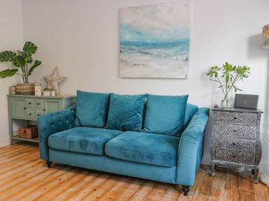 A teal sofa with three cushions between a green sideboard with plants and decorative items and a patterned chest with a vase of flowers in a room at The Lazy Seal in Spittal