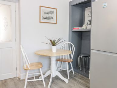 A small dining area with a round wooden table and two chairs a plant on the table a fridge and built in shelves with books and stools at The Lazy Seal in Spittal