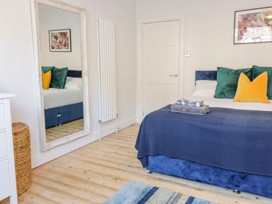 A bedroom with a blue bed, green and yellow pillows, folded towels on the bed, a large ornate mirror leaning against the wall, a wicker basket, and a framed floral picture on the wall at The Lazy Seal in Spittal