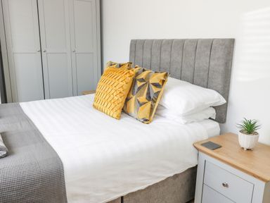 A bedroom with a bed with mustard and grey pillows a grey headboard a bedside table with a small plant and a wardrobe in the background at The Lazy Seal in Spittal